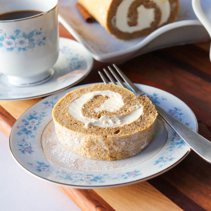 Low-carb keto pumpkin roll slice with cream cheese filling on vintage blue floral china, served with coffee and a full pumpkin roll on a wooden board in the background.
