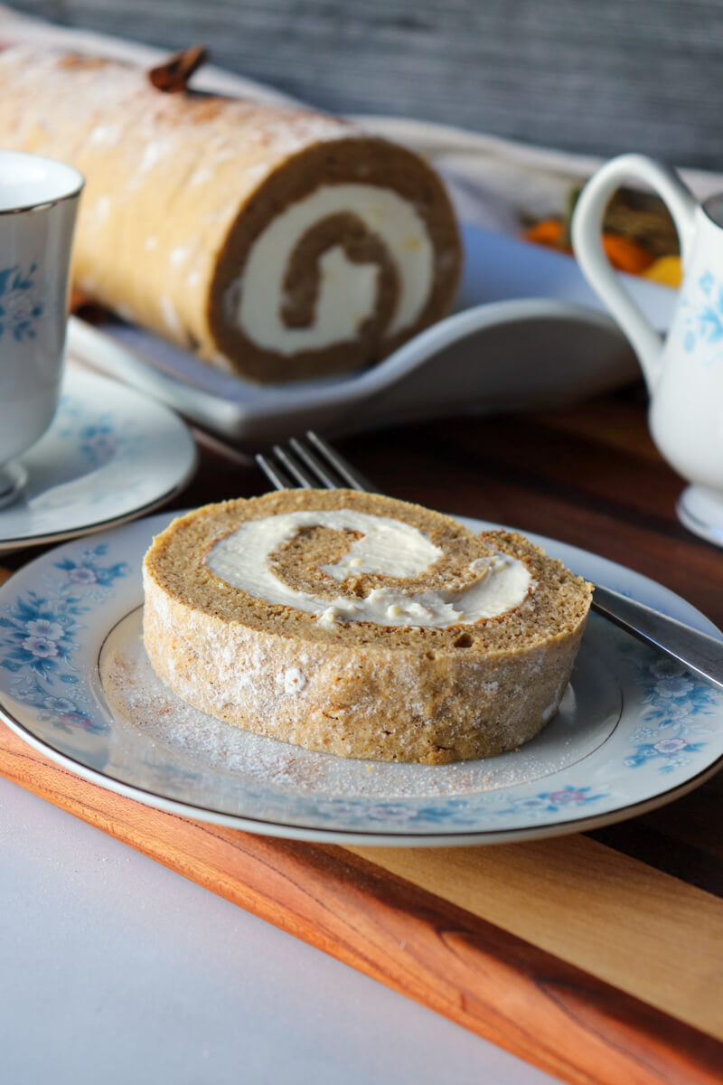 Front view of a piece of low carb pumpkin roll on a vintage china plate with blue flowers, partially eaten, with a fork on the plate, next to a cup of coffee and the rest of the roll
