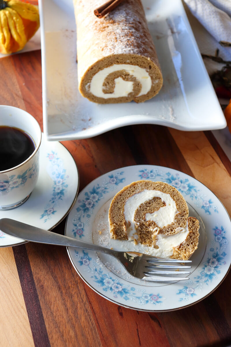 Overhead view of a piece of low carb pumpkin roll on a vintage china plate with blue flowers, partially eaten, with a fork on the plate, next to a cup of coffee and the rest of the roll