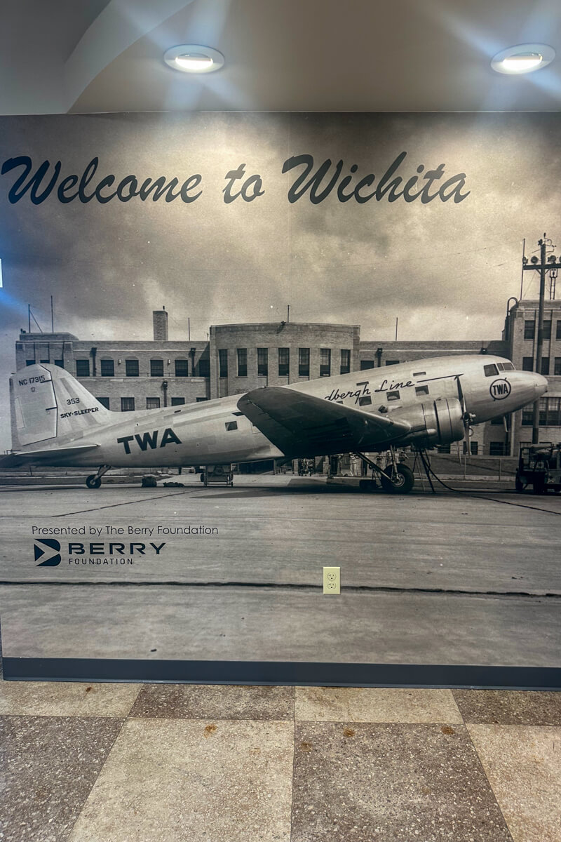 A black and white photo mural of a TWA airplane in front of the Kansas Aviation Museum building. Text: Welcome to Wichita