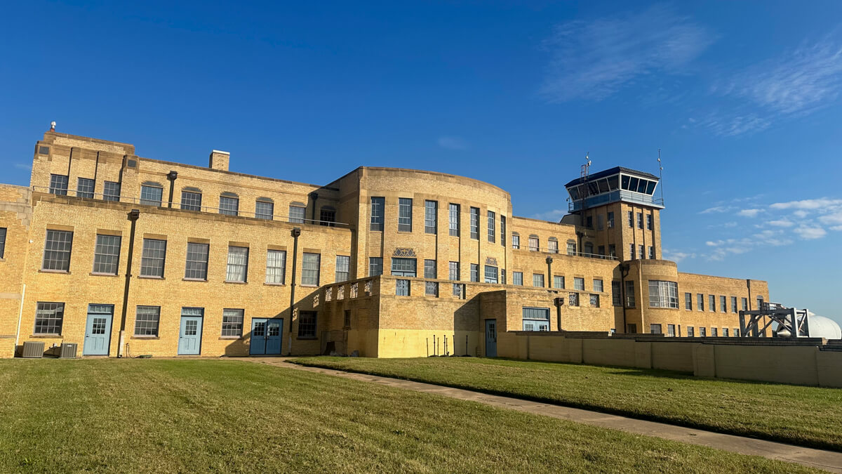 Front view of a grassy area in front of the tan art deco building with many windows that is the Kansas Aviation Museum