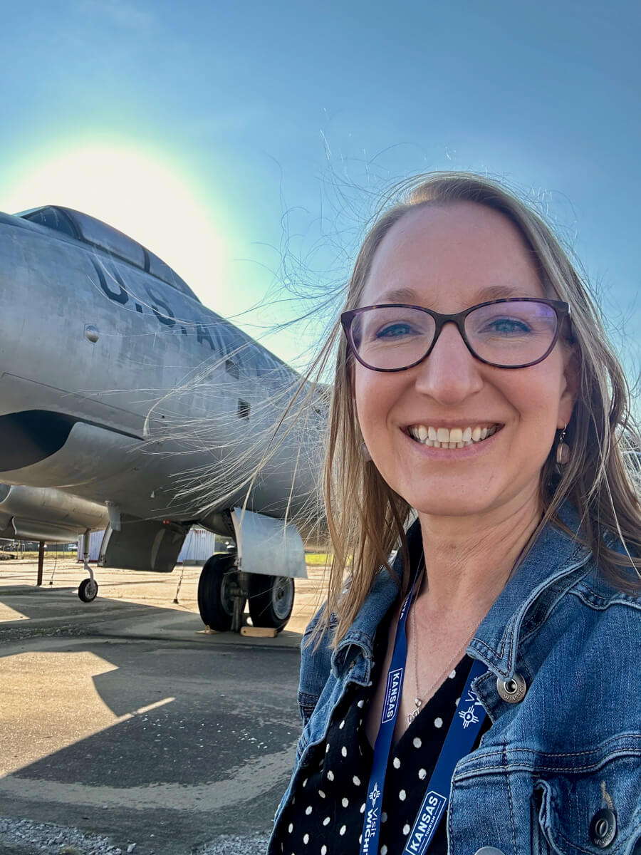 A blond woman in glasses and a jean jacket smiling in front of a large silver airplane with stenciled U.S. Air Force on the side, with blue sky in the background