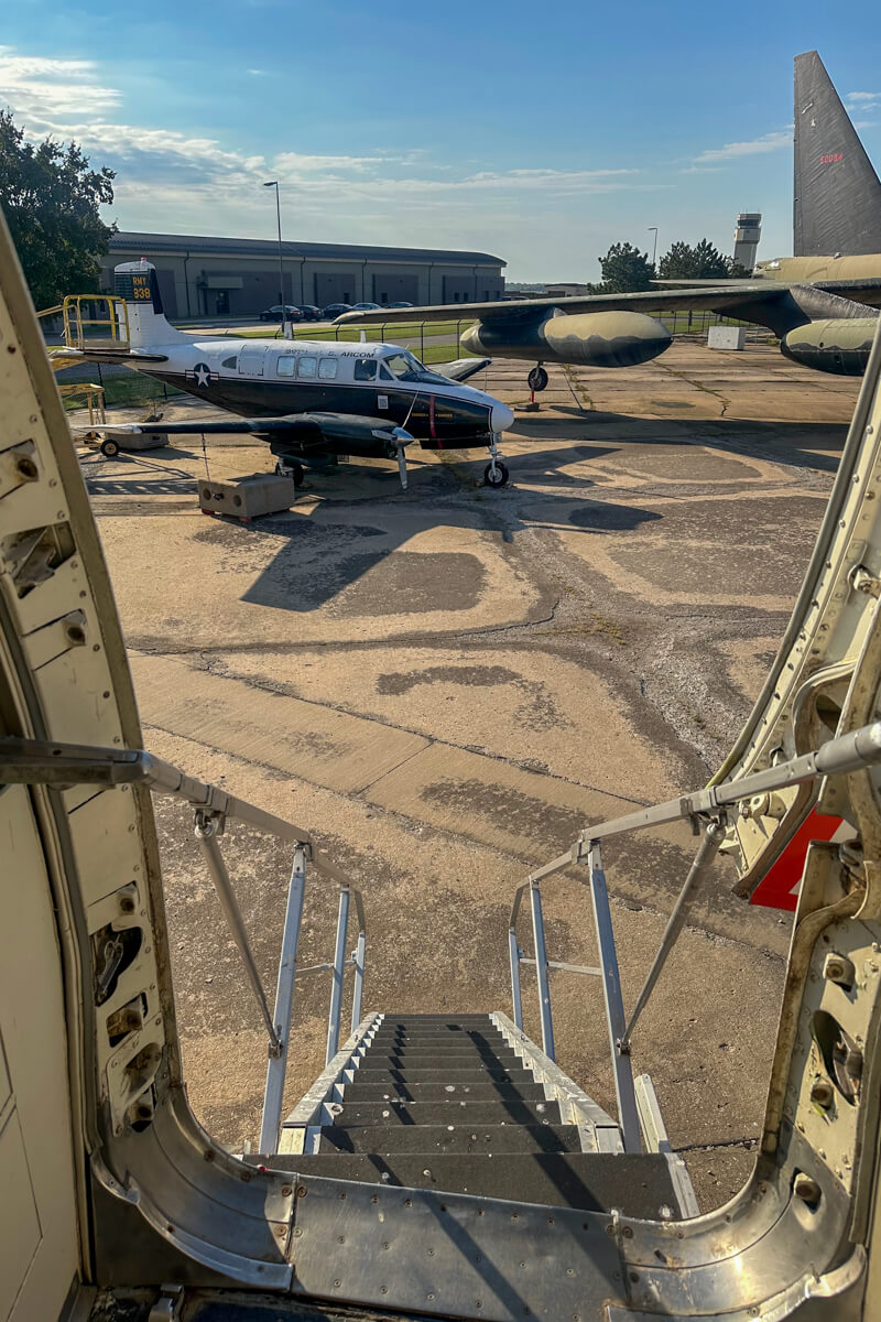 View from inside an airplane looking down the stairs with metal hand rails to the concrete with other airplanes in the distance