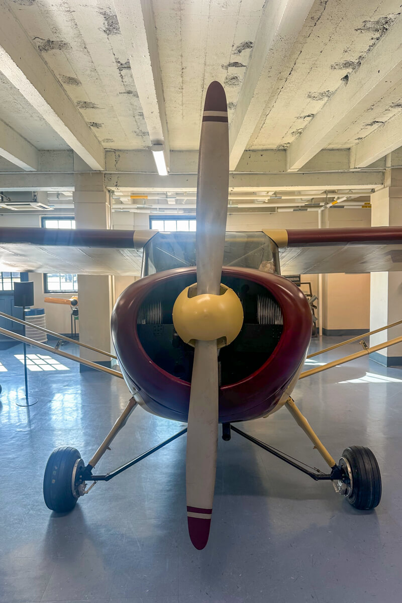 Front view of maroon and cream small airplane with propeller inside the museum room display