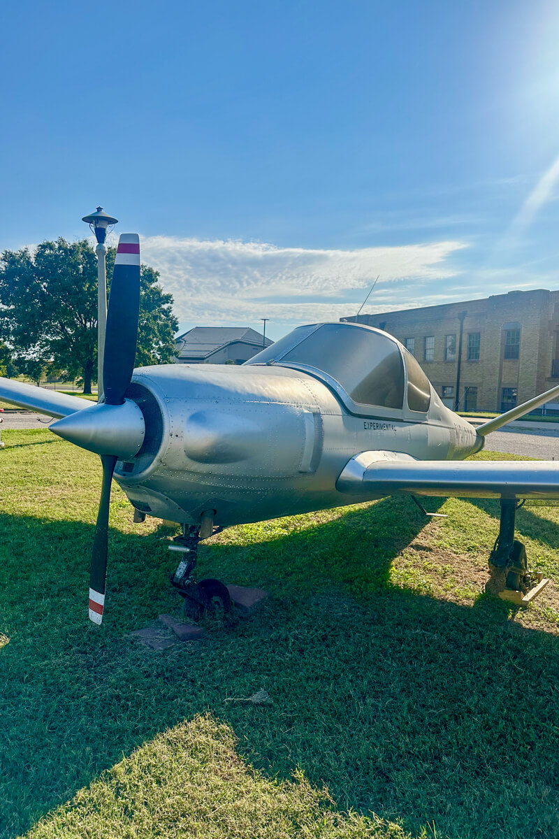 Front view of a small silver airplane with propeller on a grassy area