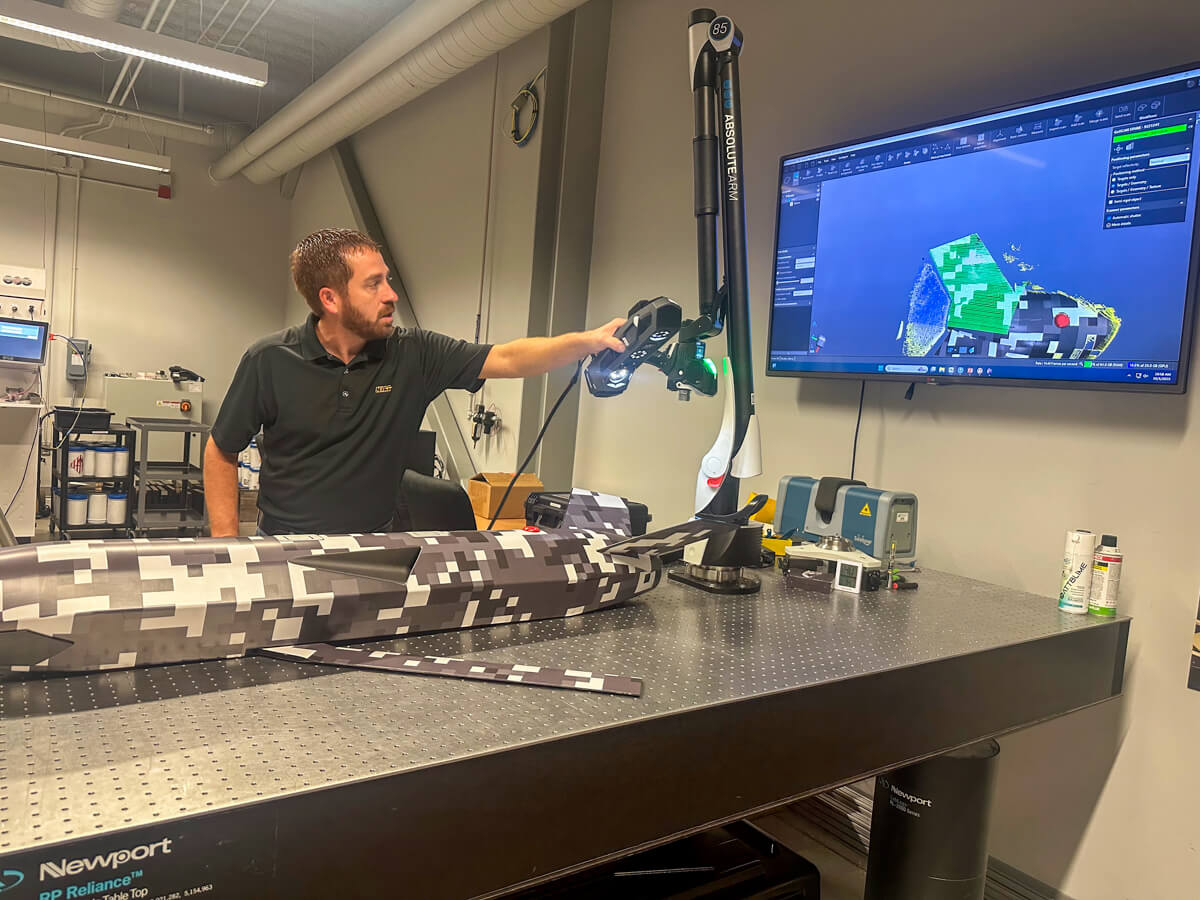 Dark red haired man with a beard demonstrating a robotic arm and scan of items displayed on a screen