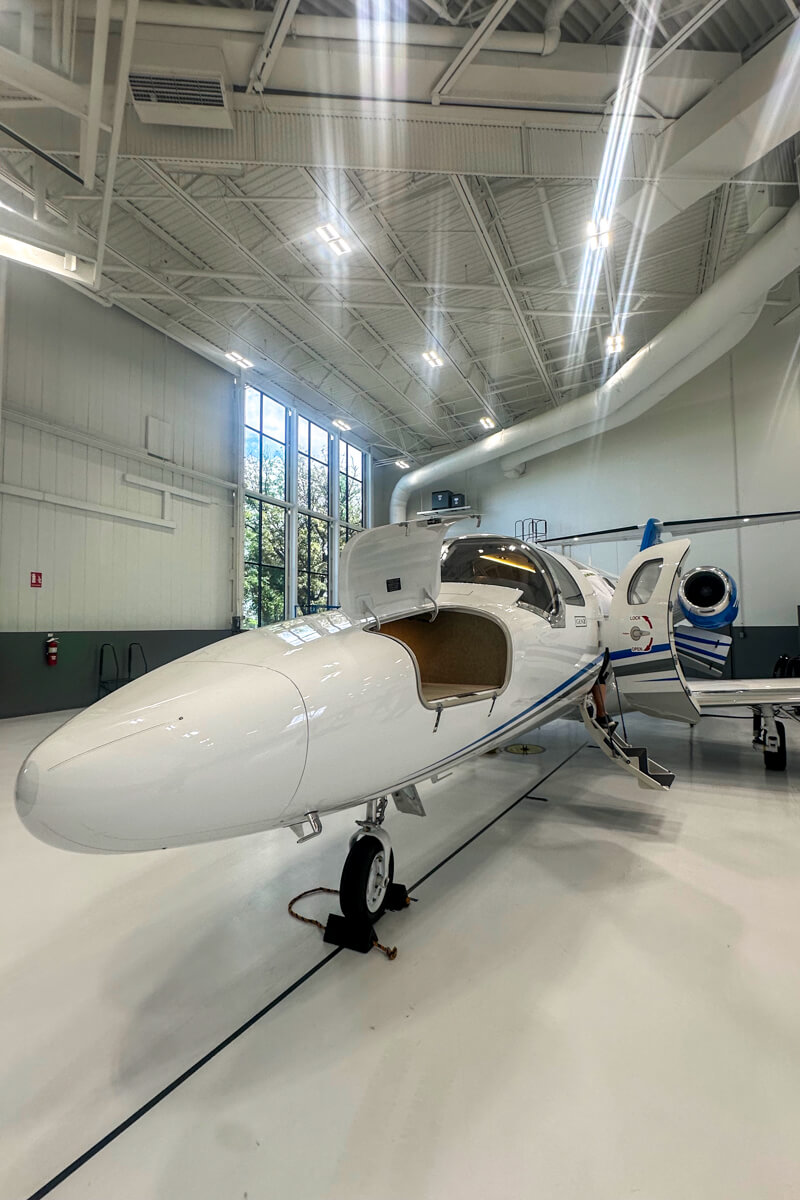 Front view of a small white private jet with the side door and front storage area open, in a large white room/hangar.
