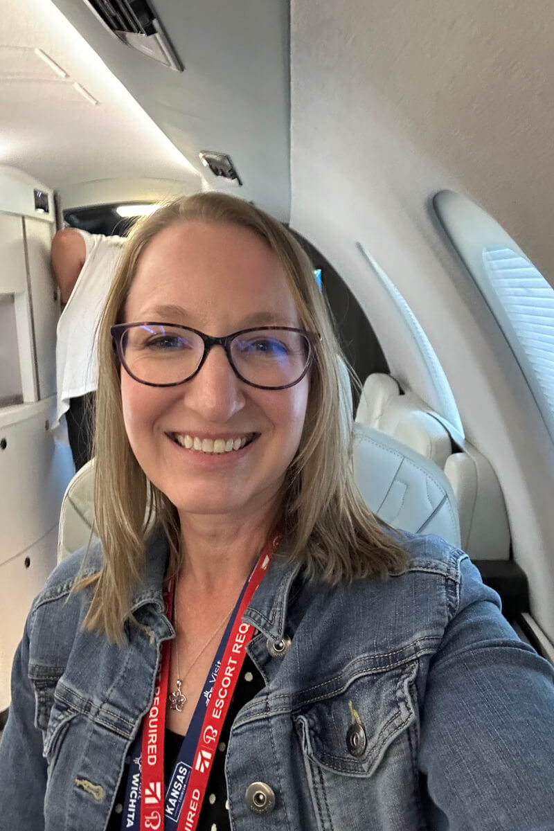 A blond woman smiling at the camera in classes and a jean jacket inside the white cabin of a private jet