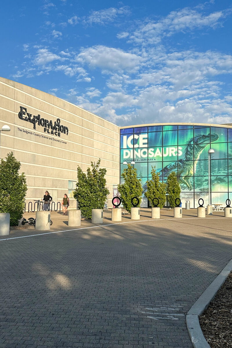 Front of the gray stone Exploration Place building with a large blue green Ice Dinosaurs display on one wall