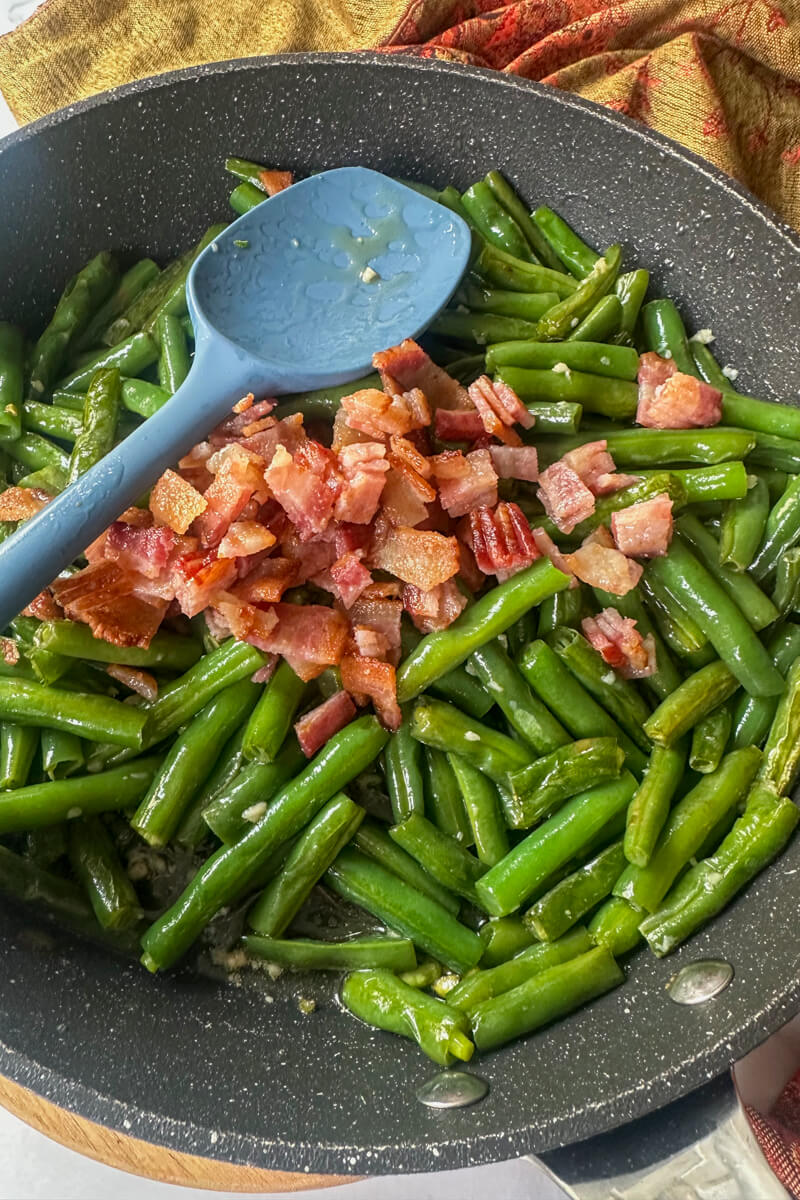 Black skillet of green beans with pieces of bacon in the center with a silicone blue spoon