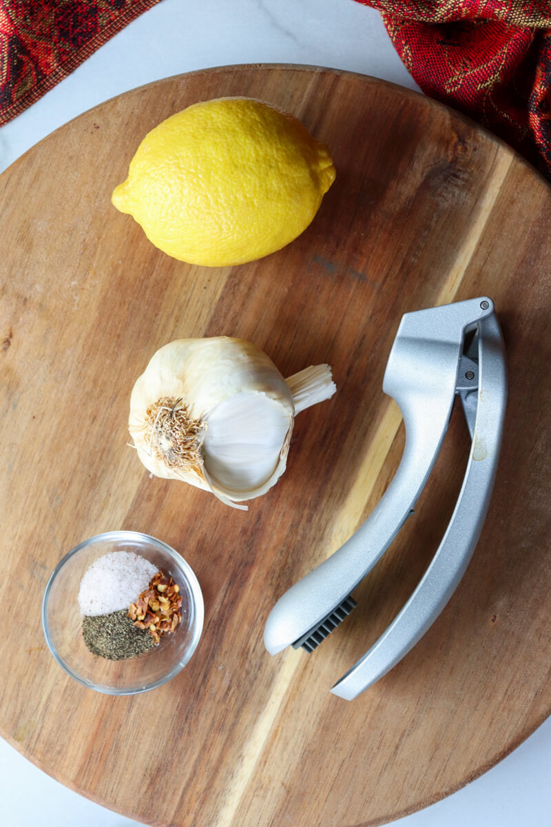 Overhead view of a head of garlic, lemon, small glass bowl of salt, pepper, and red pepper with a garlic press on a wooden circle board