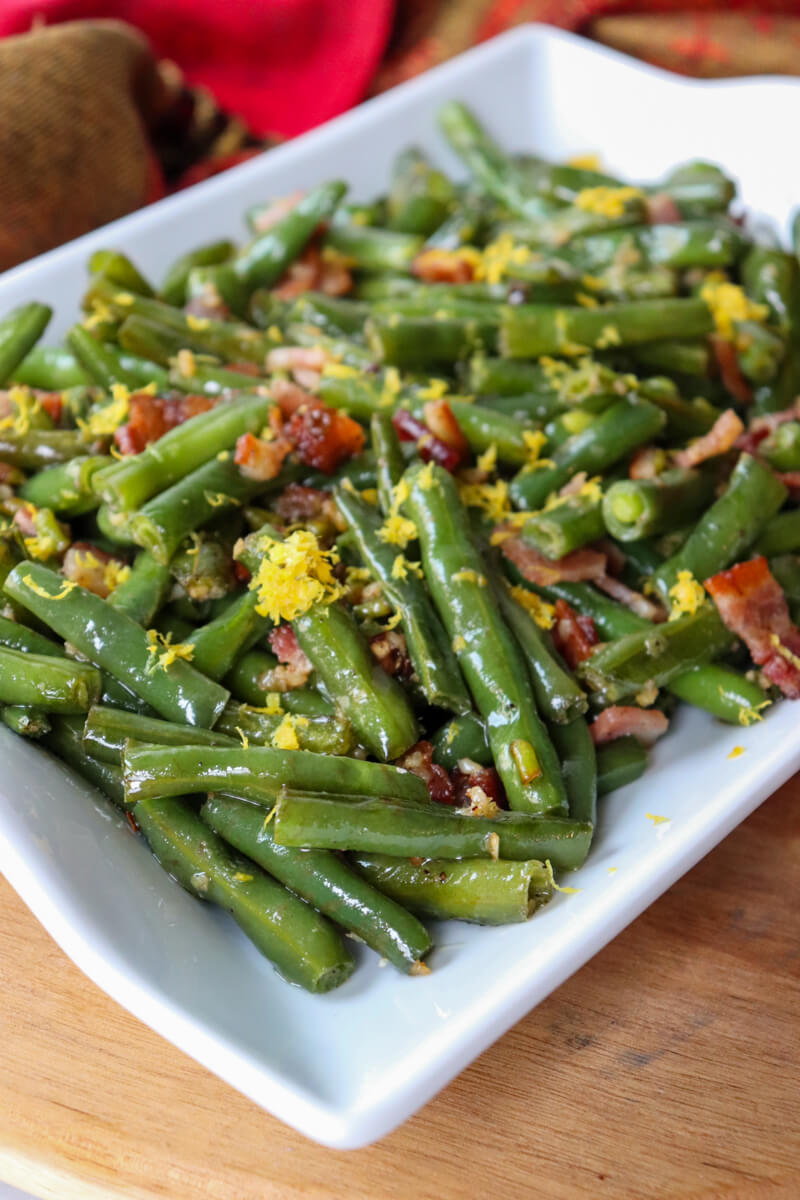Front overhead side view of green beans with bacon on a white serving dish with lemon zest