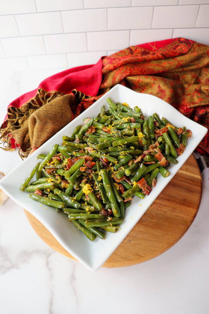 Overhead side view of green beans with bacon on a white serving dish with lemon zest