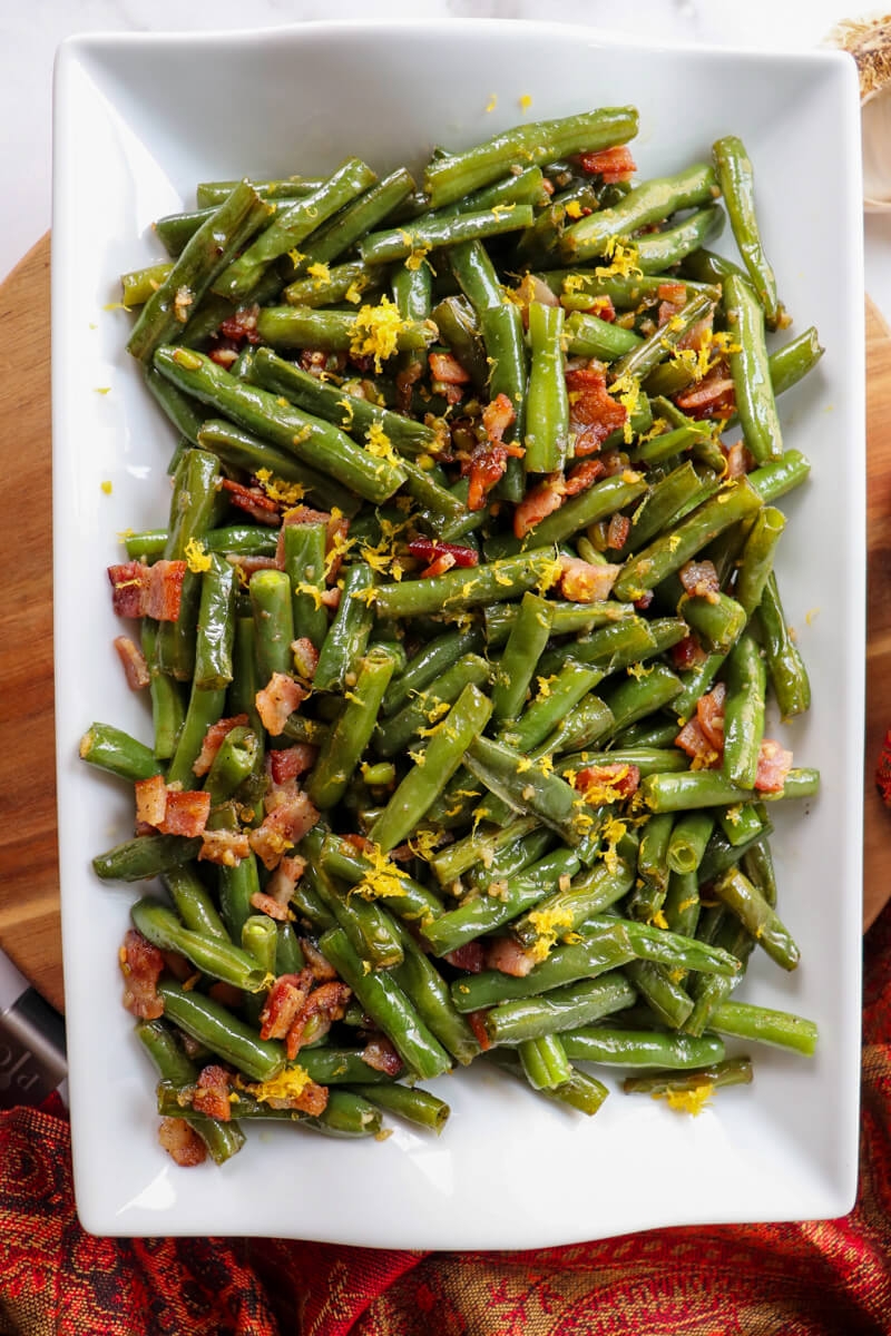 Overhead view of a white serving dish of sauteed garlic butter green beans with bacon and lemon zest
