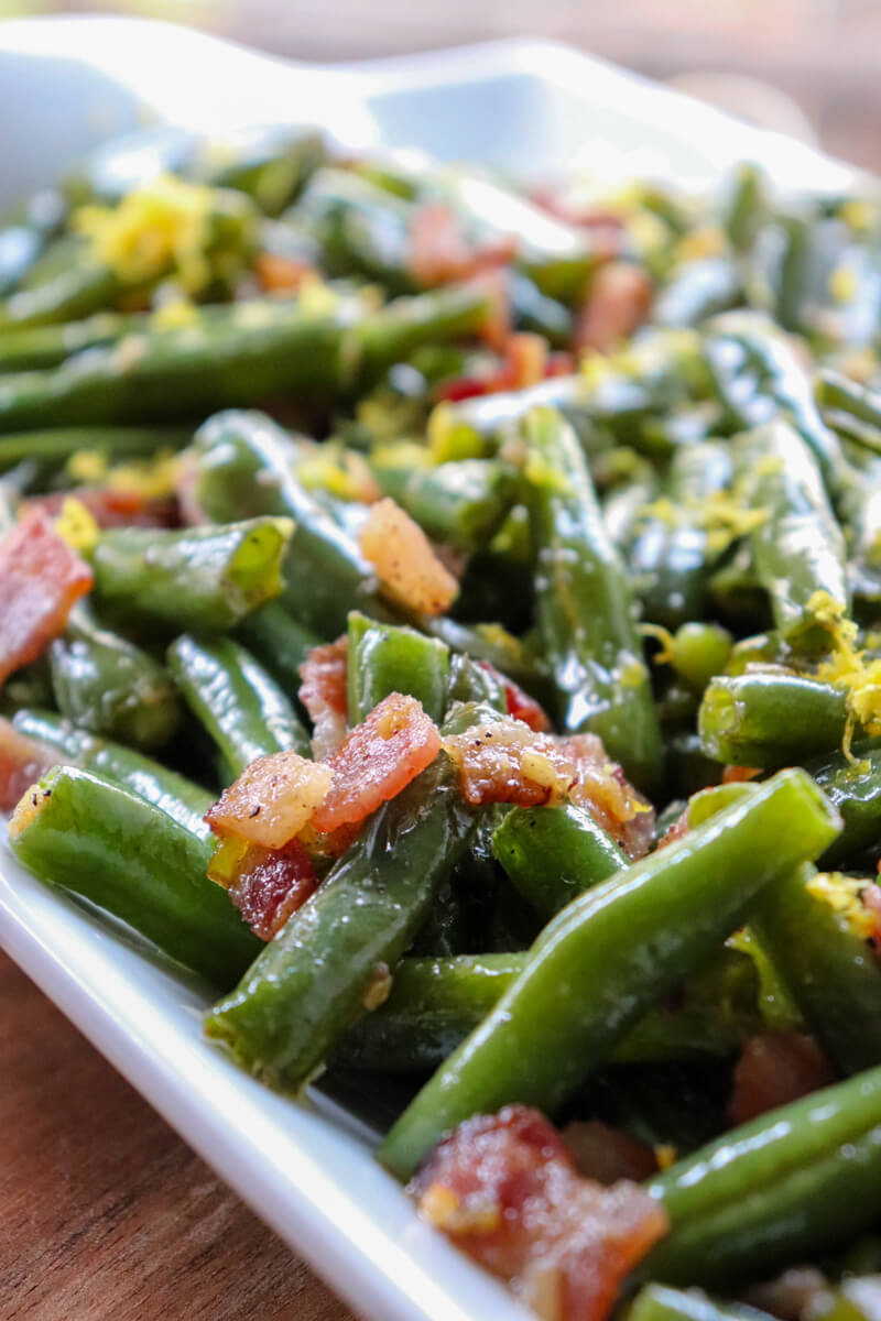 Close up view of green beans with bacon on a white serving dish with lemon zest