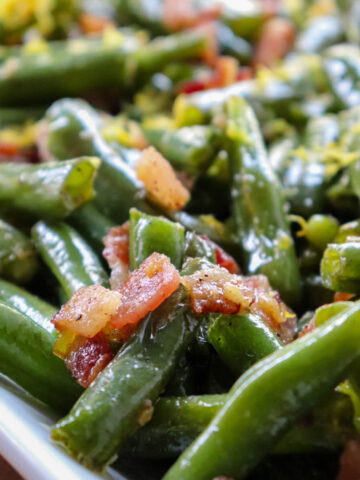 Close up view of green beans with bacon on a white serving dish with lemon zest