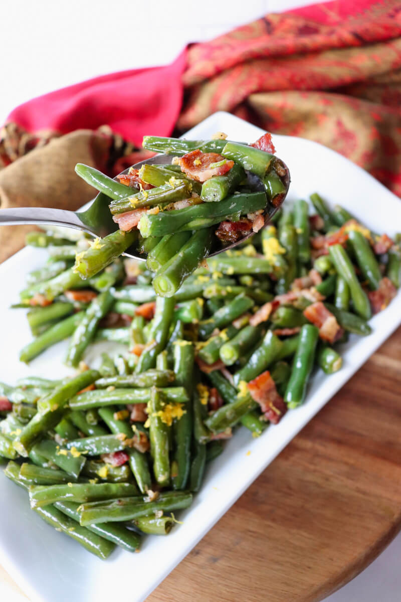 Overhead side view of green beans with bacon on a white serving dish with lemon zest with a spoonful lifting up a serving