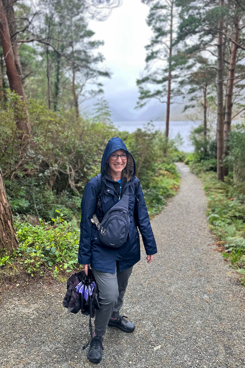 A woman in a navy raincoat on a path lined by greenery and trees toward the lake