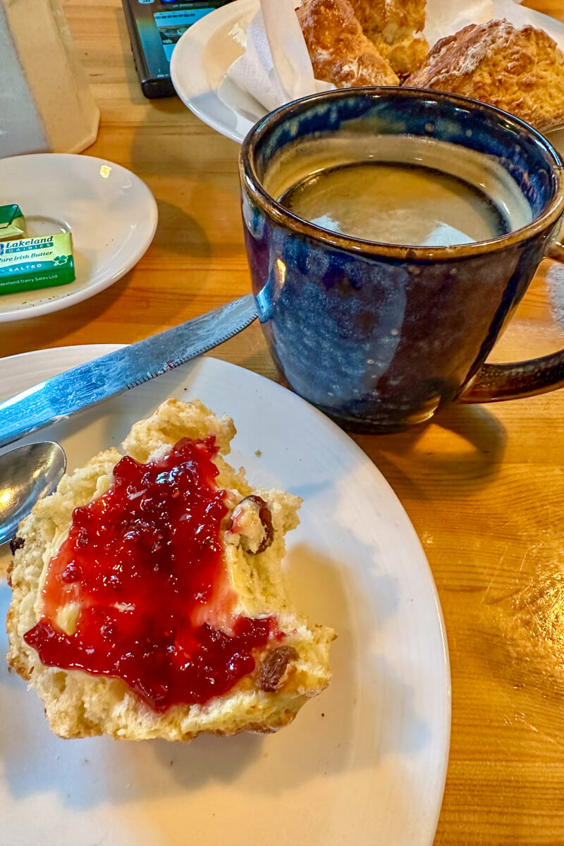 A scone with clotted cream and raspberry jam next to a navy mug of coffee (Americano)
