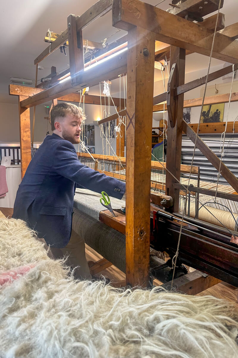A man in a blue tweed coat sitting at and working a traditional wool loom making Donegal tweed.