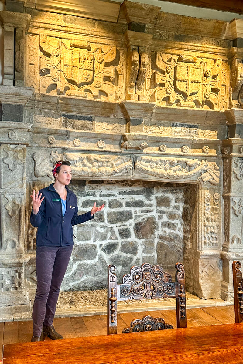 Emma in jeans and a navy jacket stands and speaks in front of the famous stone fireplace at Donegal Castle