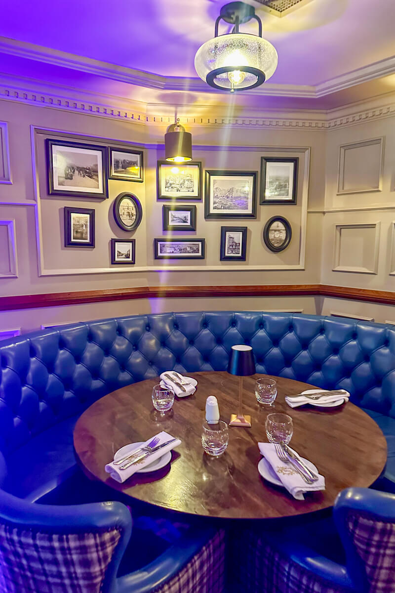 A round wooden table in a cozy nook of the Market House Restaurant. Blue padded seats and framed pictures on the wall.