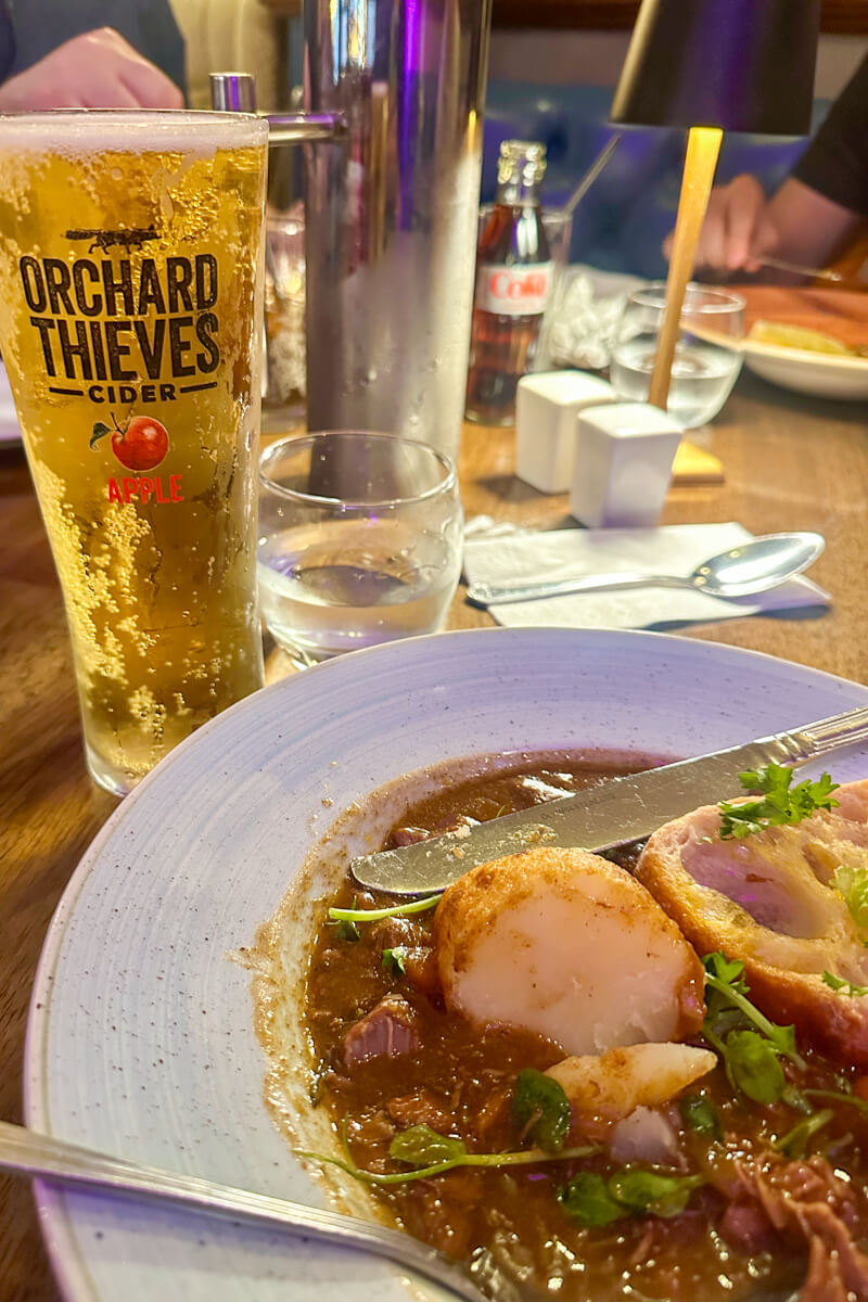 A plate of beef and Guinness stew on a white plate next to a pint of Orchard Thieves cider.