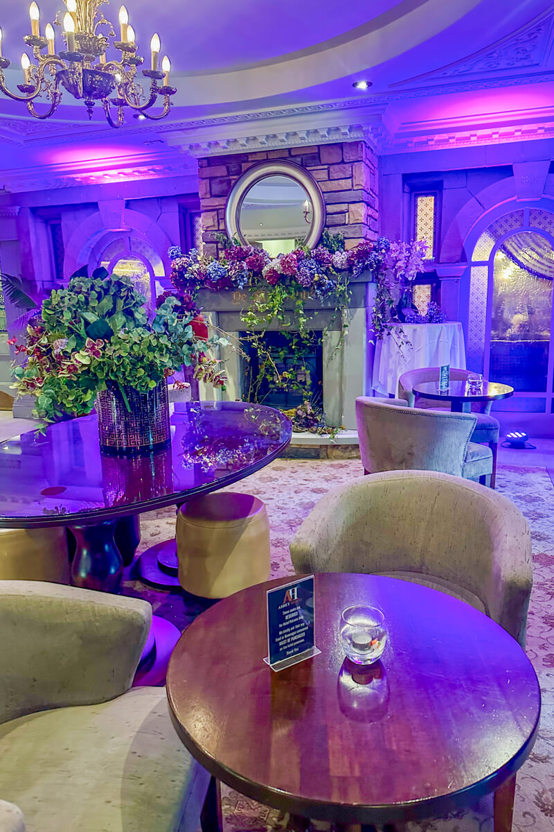 Inside lighting at the cosy Abbey Hotel lobby with a chandelier, wooden tables, and comfy tan padded chairs