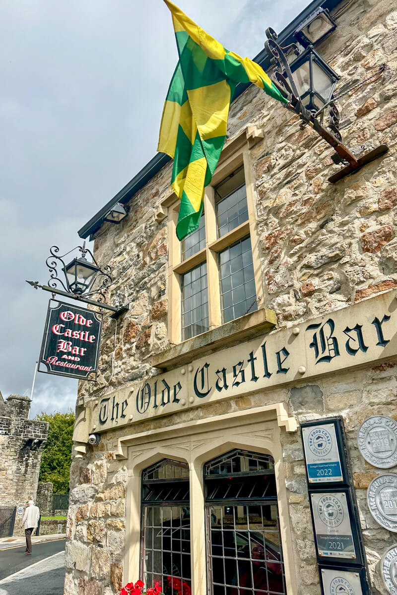 A front view of the stone walled side of The Olde Castle Bar with a green and yellow flag hanging down as well as a cast iron sign for the bar.