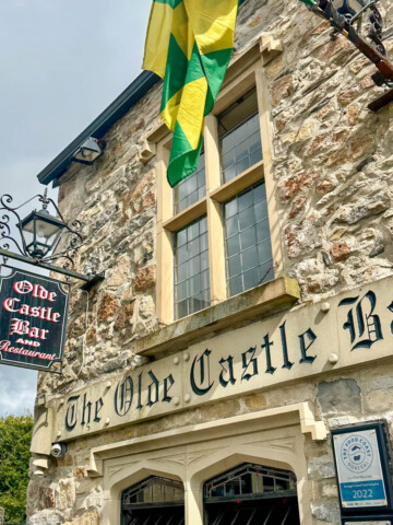A front view of the stone walled side of The Olde Castle Bar with a green and yellow flag hanging down as well as a cast iron sign for the bar.