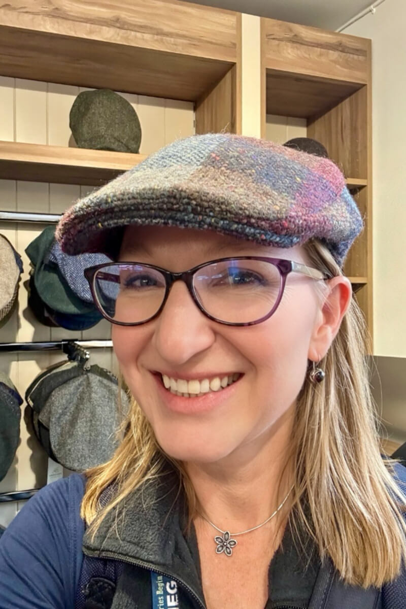 A woman with straight dirty blond hair and classes looking at the camera wearing a patchwork flatcap Donegal tweed hat