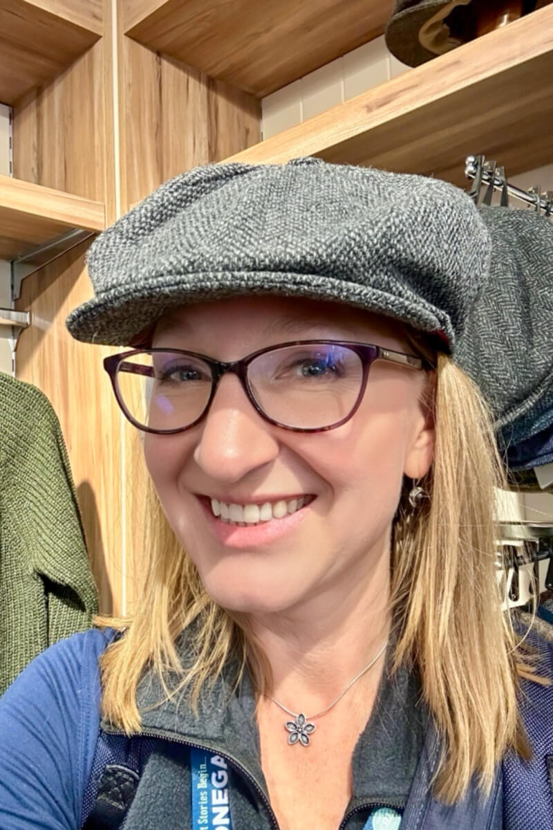 A woman with straight dirty blond hair and classes looking at the camera wearing a gray Donegal tweed newsboy hat