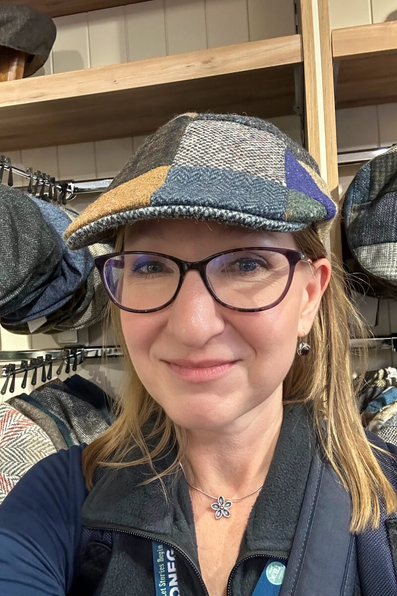 A woman with straight dirty blond hair and classes looking at the camera wearing a patchwork duckbill Donegal tweed hat