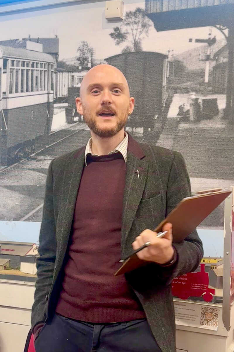 A bald man with a small moustache and beard in a maroon sweater and brown Donegal tweed coat speaking in front of train posters