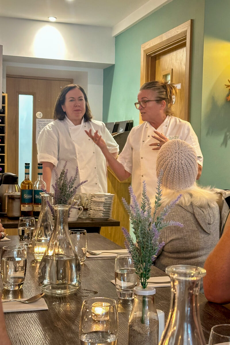 The two chefs in white chef coats at the head of the table speaking and making hand motions.