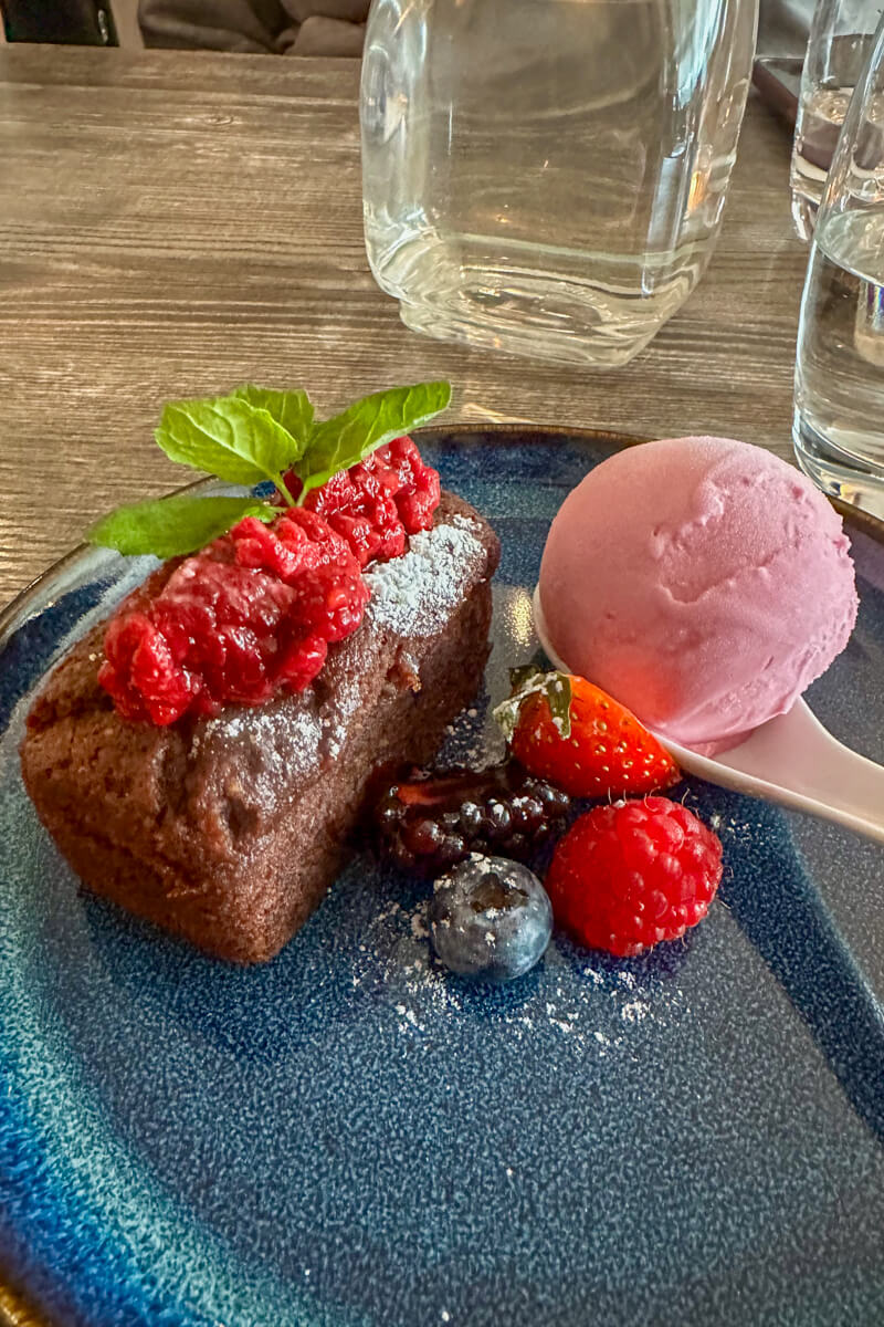 Front view of a gluten free brownie with raspberries on top, blueberries, blackberries, etc. next to it, and a scoop of raspberry sorbet on a dark blue plate.