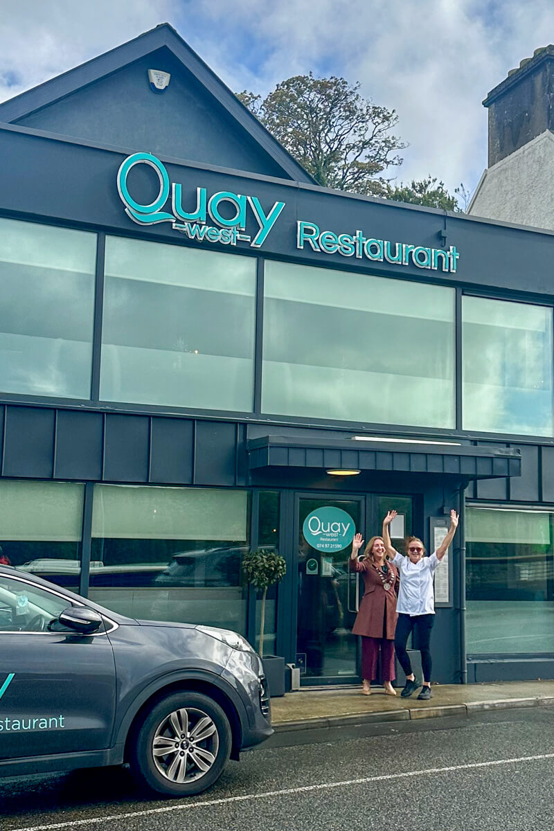 Two women waving from the front of the Quay Restaurant. It's dark gray colored with large windows.