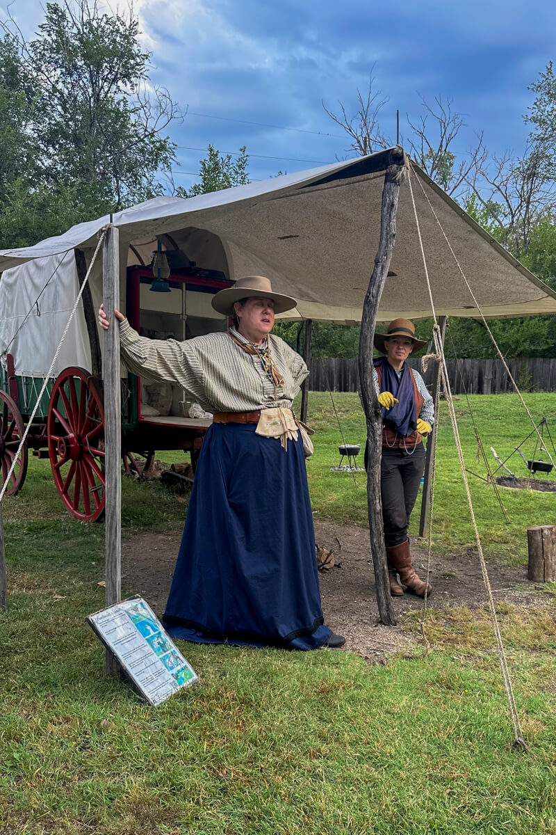 Cowtown Wichita actors at a cowboy camp over a picture of a covered wagon glamping area with a picnic table.