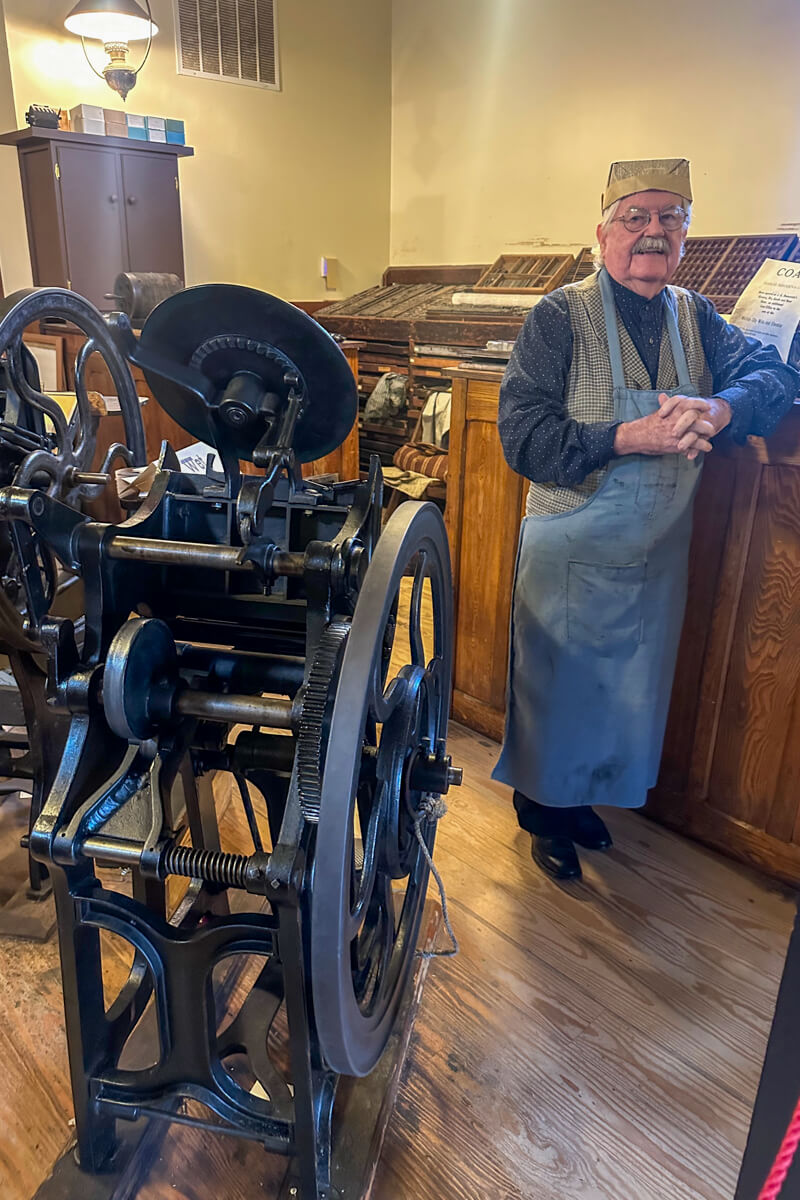 A vintage printing press with a man in vintage printer's uniform standing next to it