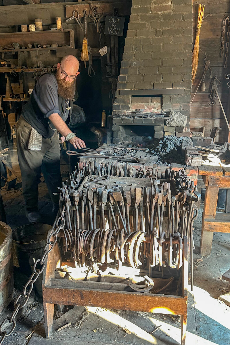 The blacksmith leaning over the work area and fire next to the bellows with metal tools in front