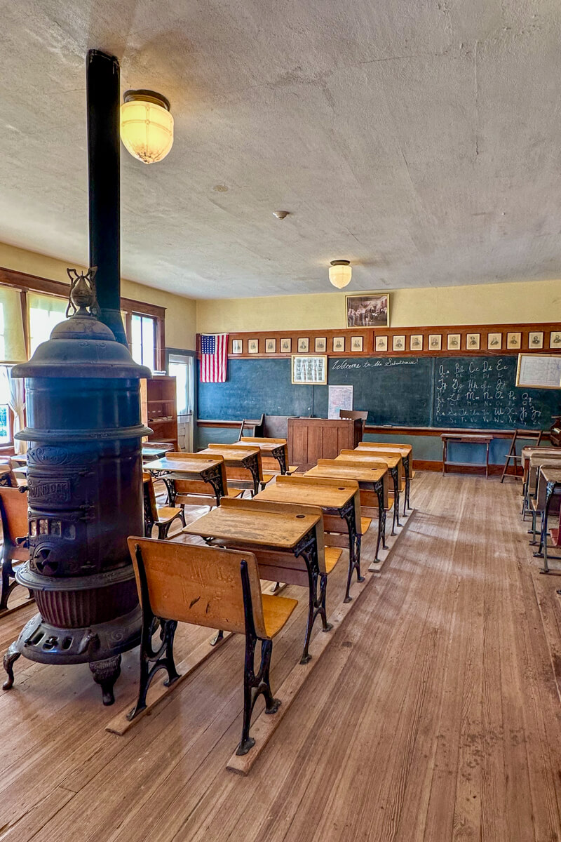 The view inside a one room school house with the wood stove, lines of desks, and chalkboard at the front of the room