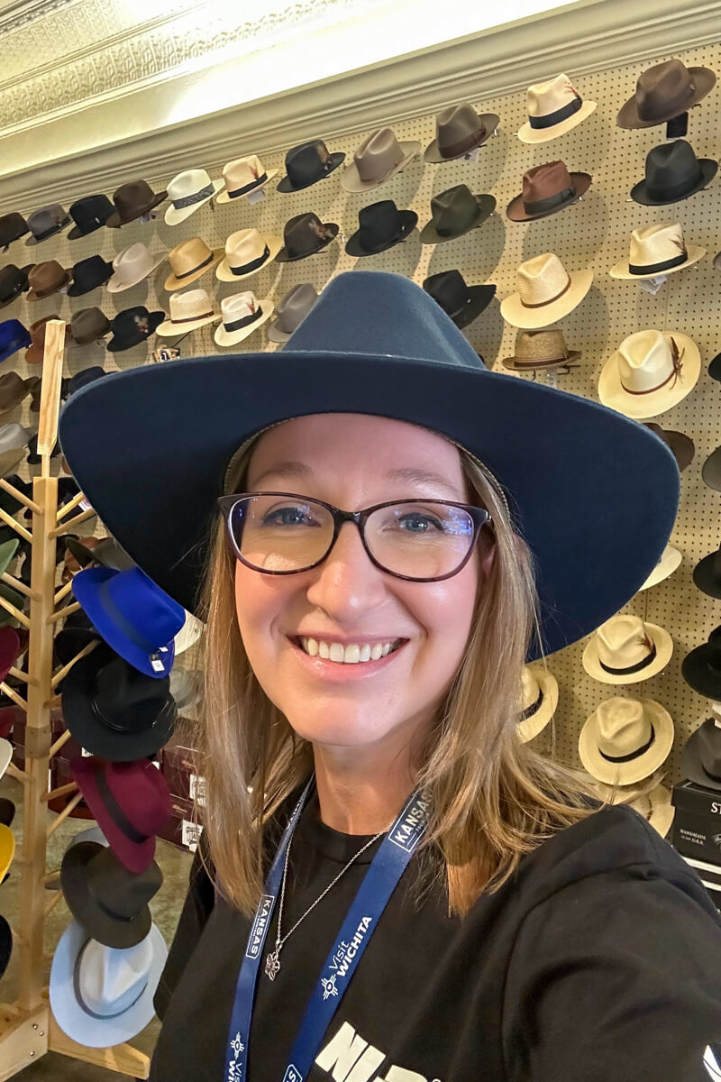 a blond woman in a navy hat, black tshirt and glasses in front of a wall display of different hats