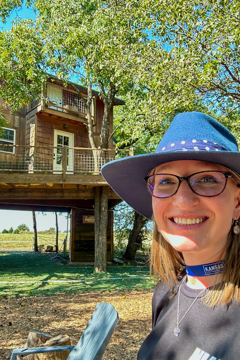 A blond woman in a navy hat, black tshirt and glasses standing in front of a two story cabin tree house