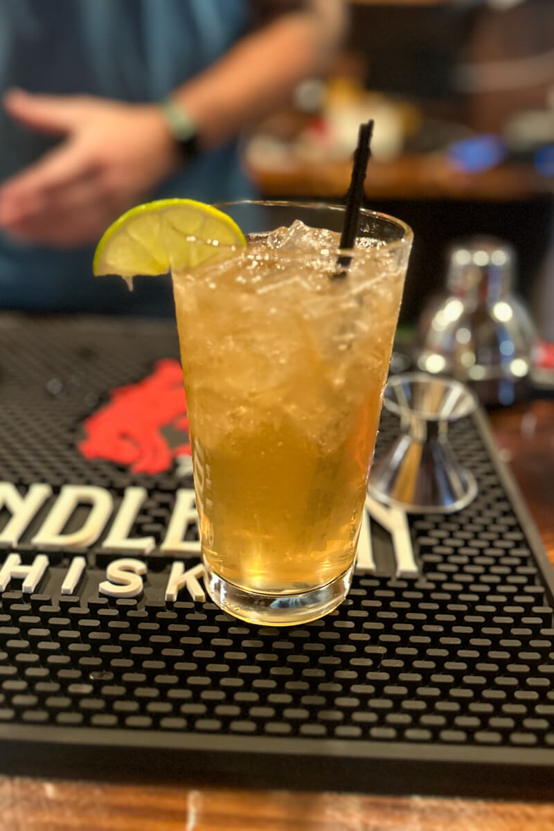 A tall golden colored drink in a glass with a black straw and a lime wedge