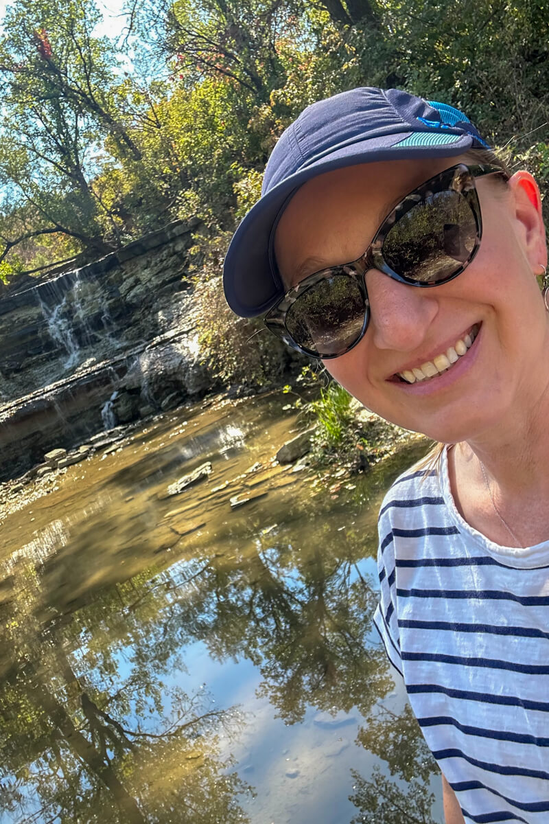 A view of stones and calm water looking up towards a small waterfall surrounded by trees with a woman in sunglasses, a navy cap, and a white/navy striped t-shirt in front
