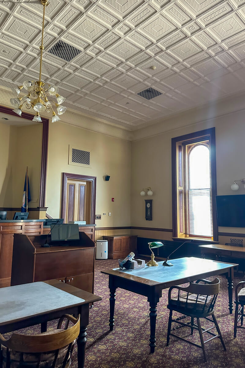 The main courtroom at the courthouse with tall ceiling, light coming in the wooden arched window, and wooden tables, etc.