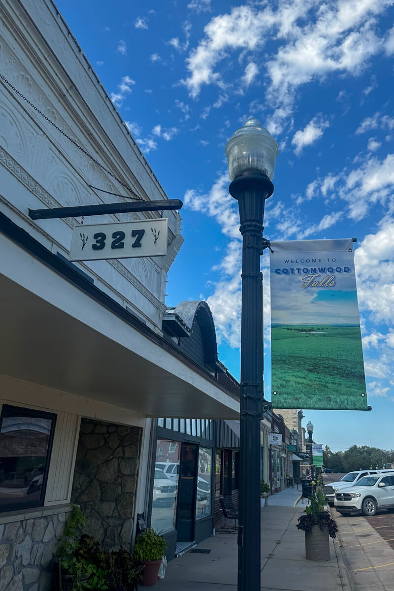 A main street store front view with a vintage style lamp pole and sign that says "Welcome to Cottonwood Falls".