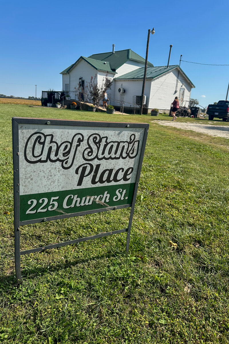 A green and white sign that says "Chef Stan's Place" in front of a white prairie farmhouse style building