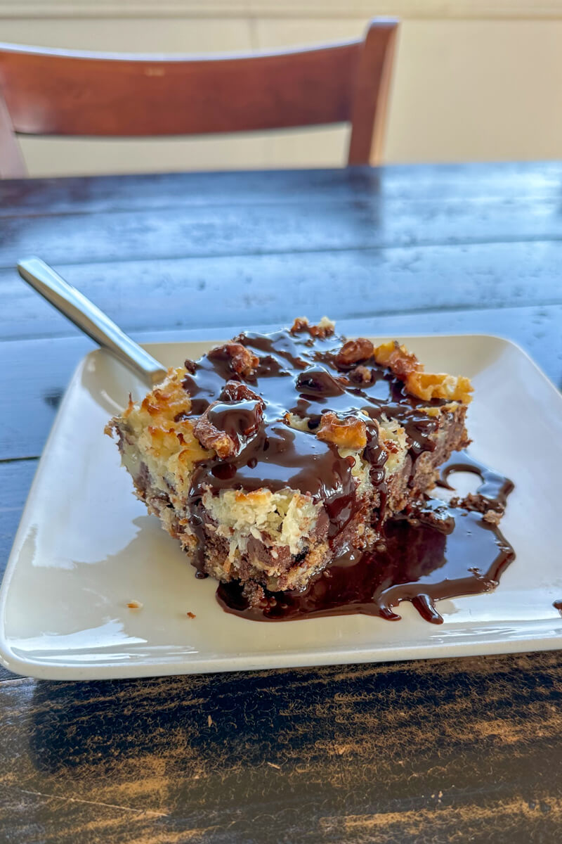 A giant magic cookie bar covered in chocolate sauce on a white plate on a wooden table
