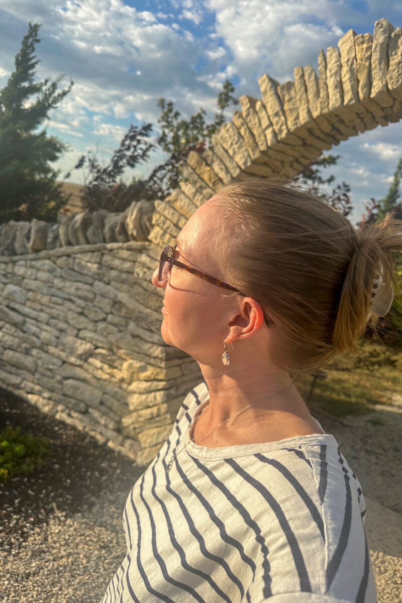 A blond woman with hair pulled back and glasses looking back towards the sun with a stone archway in the background